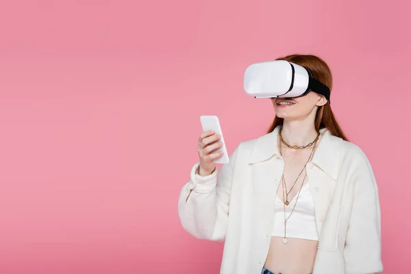 Mujer elegante sonriente en auriculares de realidad virtual usando teléfono celular aislado en rosa - foto de stock