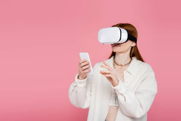 Stylish redhead woman in virtual reality headset using mobile phone isolated on pink — Stock Photo