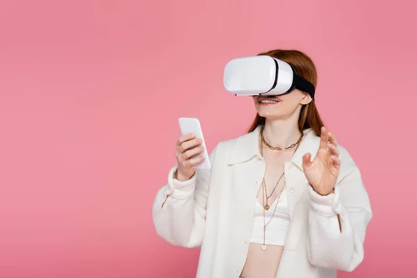 Mulher positiva em jogos de jaqueta no fone de ouvido vr e usando smartphone isolado em rosa — Fotografia de Stock