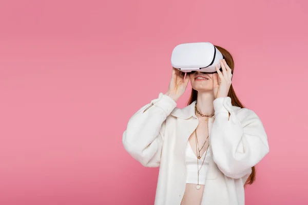 Sorrindo mulher de cabelos vermelhos em jaqueta e colares jogos em vr fone de ouvido isolado em rosa — Fotografia de Stock