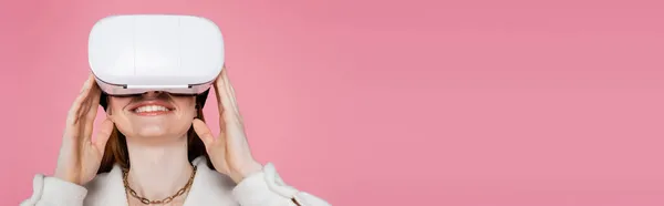 Mujer de pelo rojo positivo en chaqueta y collar usando auriculares vr aislados en rosa, bandera - foto de stock