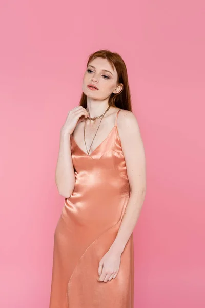 Elegante mujer pelirroja en vestido mirando hacia otro lado aislado en rosa - foto de stock