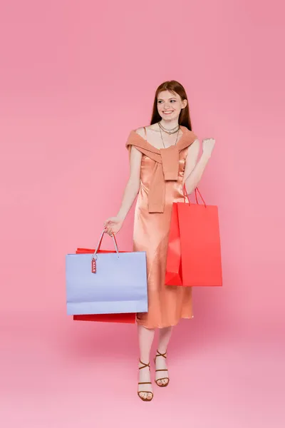 Mujer sonriente en vestido de seda sosteniendo bolsas de compras con etiqueta de precio sobre fondo rosa - foto de stock