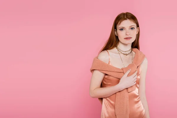 Red haired woman in peach silk dress and sweater looking at camera isolated on pink — Stock Photo