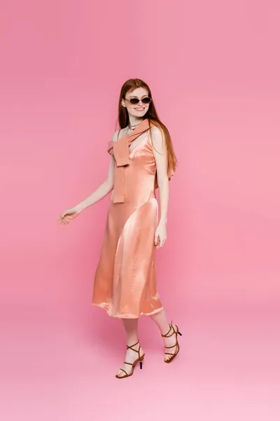 Stylish woman in silk dress and sunglasses walking on pink background — Stock Photo