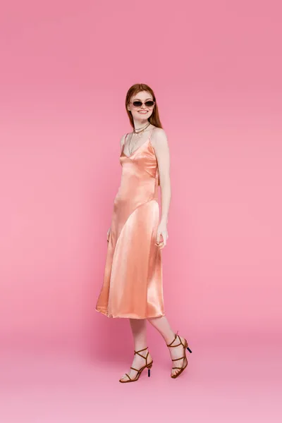 Улыбающаяся женщина в шелковом платье и солнцезащитных очках ходит на розовом фоне — стоковое фото