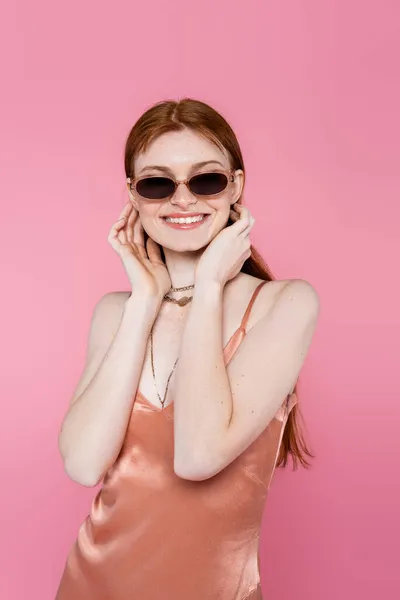 Усміхнена веснянка жінка в сонцезахисних окулярах і персиковій сукні ізольована на рожевому — стокове фото