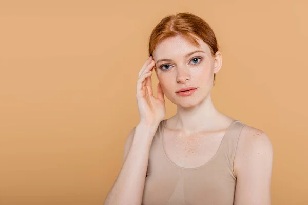 Portrait of young freckled woman touching face and looking at camera isolated on beige — Stock Photo