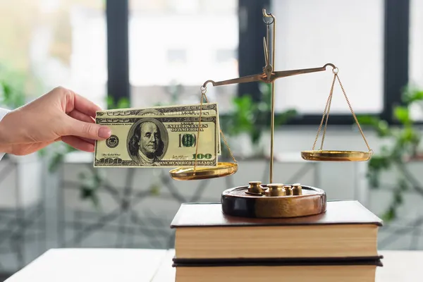 KYIV, UKRAINE - SEPTEMBER 22, 2021: cropped view of woman holding dollars near justice scales on books in office, anti-corruption concept — Stock Photo
