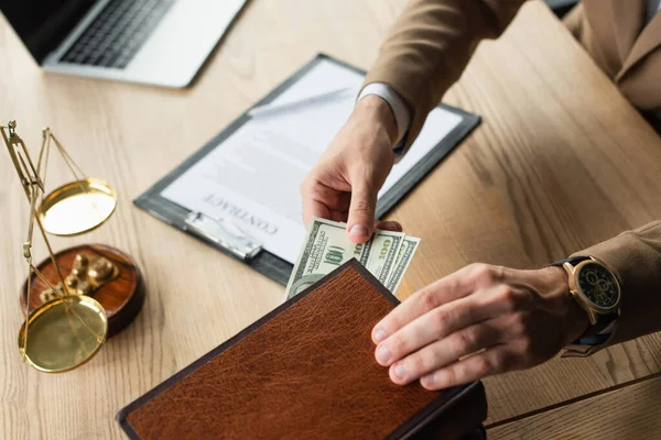 Cropped view of lawyer hiding money in codex book near justice scales and blurred contract, anti-corruption concept — Stock Photo