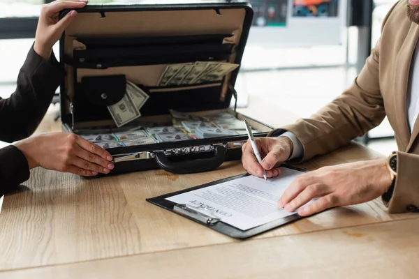 Vue partielle de l'homme d'affaires signant contrat près de la femme avec des dollars dans la mallette, concept anti-corruption — Photo de stock