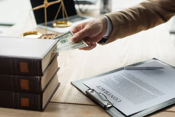 Abgeschnittene Ansicht des Anwalts, der Dollars in ein Buch in der Nähe von Verträgen und verschwommenen Gerechtigkeitsmaßstäben einbringt, Anti-Korruptions-Konzept — Stockfoto