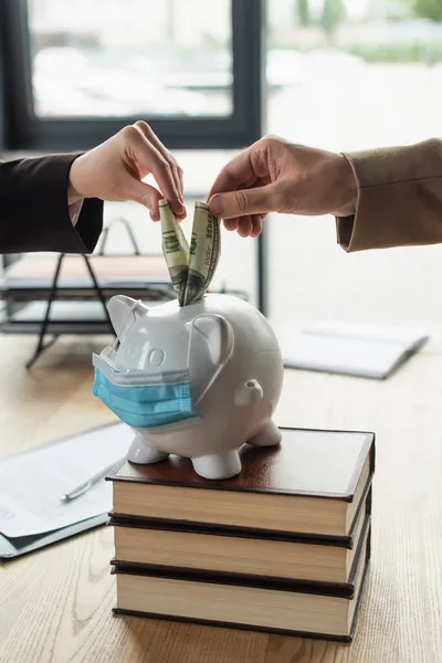 Cropped view of lawyer and client putting dollars into piggy bank in medical mask, anti-corruption concept — Stock Photo