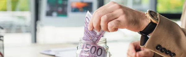 Частичный взгляд бизнесмена возле стеклянной банки с банкнотами евро, антикоррупционная концепция, баннер — стоковое фото