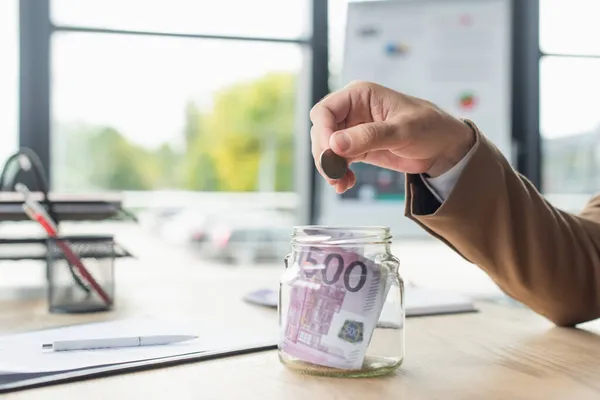 Partial view of businessman holding coin near glass jar with euro banknotes in office, anti-corruption concept — Stock Photo