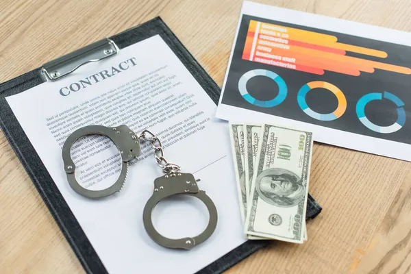 KYIV, UKRAINE - SEPTEMBER 22, 2021: top view of handcuffs and dollars near contract and infographics on desk, anti-corruption concept — Stock Photo