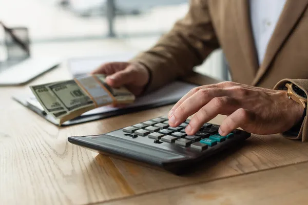 Обрізаний погляд бухгалтера, який використовує калькулятор, тримаючи доларові банкноти, антикорупційна концепція — стокове фото