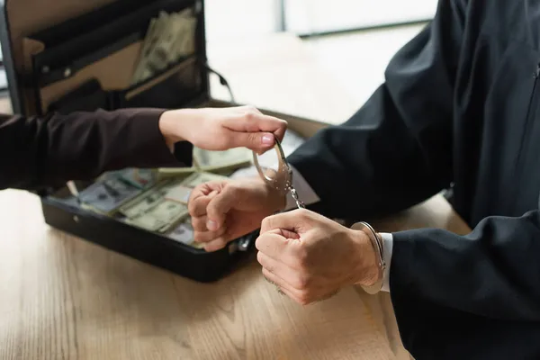 Vista recortada de la mujer que pone esposas en hombre de negocios cerca difuminado maletín con dólares, concepto anticorrupción - foto de stock