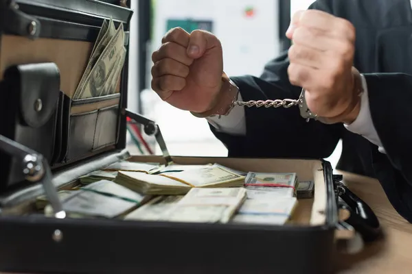 Partial view of handcuffed businessman near blurred briefcase with dollars, anti-corruption concept — Stock Photo