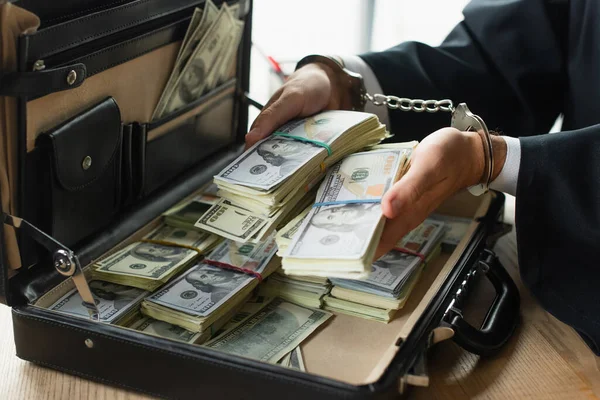 Частковий погляд бізнесмена в наручниках, що тримає долари біля портфеля, антикорупційна концепція — стокове фото