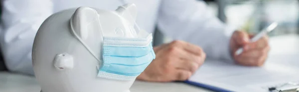 Close up view of piggy bank in medical mask near cropped doctor and clipboard on blurred background, anti-corruption concept, banner — Stock Photo