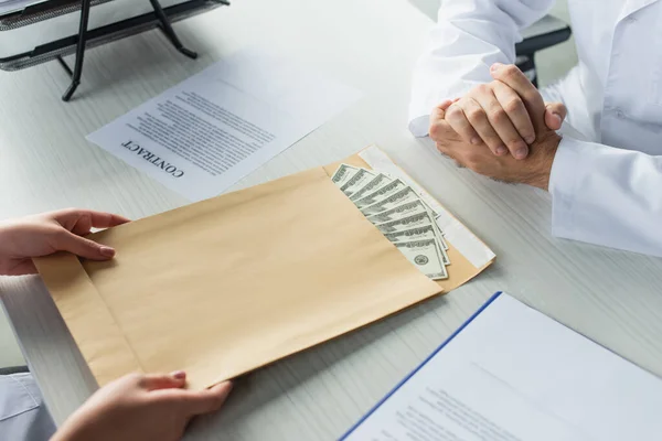 Vista recortada de la mujer sosteniendo sobre con dinero cerca del médico, concepto anticorrupción - foto de stock