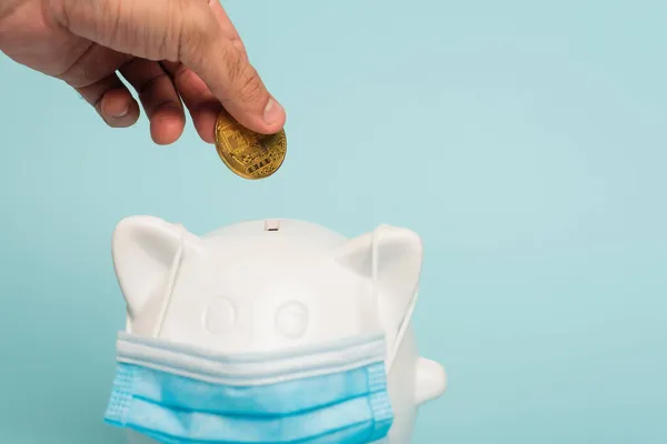 KYIV, UKRAINE - SEPTEMBER 22, 2021: cropped view of man holding bitcoin near piggy bank in medical mask isolated on blue, anti-corruption concept — Stock Photo