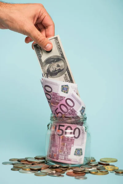 KYIV, UKRAINE - SEPTEMBER 22, 2021: cropped view of male hand near coins and glass jar with dollar and euro banknotes on blue, anti-corruption concept — Stock Photo