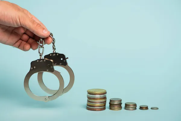 Partial view of man holding handcuffs near stacks of silver and golden coins on blue, anti-corruption concept — Stock Photo