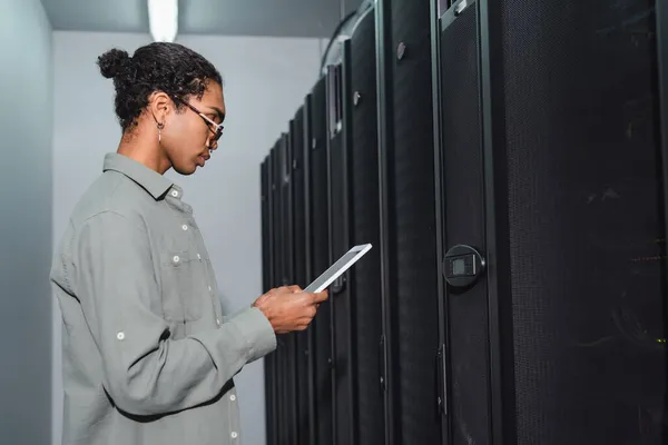 Vue latérale du programmeur afro-américain avec tablette numérique debout près des serveurs dans le centre de données — Photo de stock