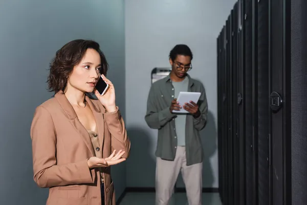 Ingeniero hablando en el teléfono móvil cerca borrosa colega afroamericano con tableta digital en el centro de datos - foto de stock