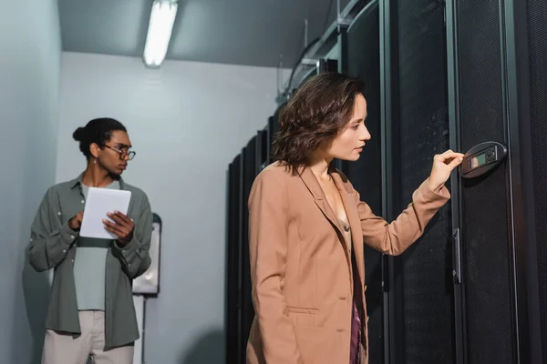 Інженер перевіряє сервер біля розмитого афроамериканського колеги в центрі обробки даних — стокове фото