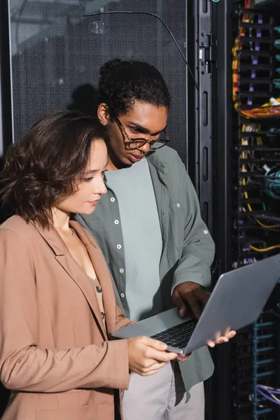 Par de ingenieros multiétnicos mirando el ordenador portátil mientras comprueba el servidor en el centro de datos - foto de stock