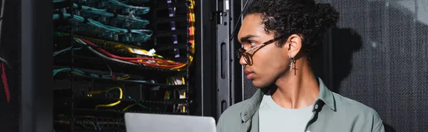 Афроамериканський інженер з ноутбуком, який дивиться на дроти на сервері під час роботи в центрі обробки даних, банер — стокове фото