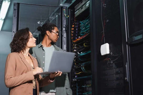 Jóvenes ingenieros interraciales con computadora portátil haciendo diagnósticos de servidor en centro de datos - foto de stock