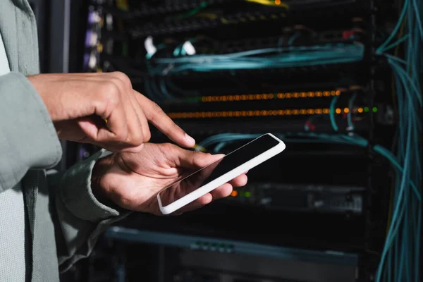 Ausgeschnittene Ansicht eines afrikanisch-amerikanischen Programmierers, der auf ein Smartphone mit leerem Bildschirm in Servernähe zeigt — Stockfoto