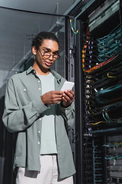Programmatore afroamericano sorridente usando il telefono cellulare mentre fa la diagnostica del server nel data center — Foto stock