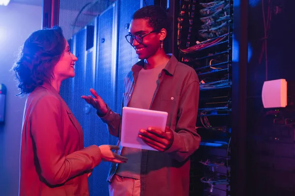 Smiling african ameican engineer with digital tablet gesturing while talking with colleague near server — Stock Photo