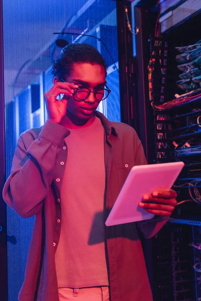 Young african american programmer adjusting eyeglasses while looking at digital tablet in data center — Stock Photo