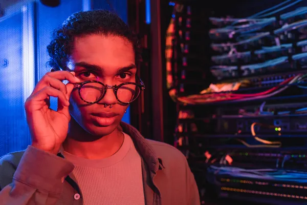 Young african american engineer touching eyeglasses while looking at camera near blurred server — Stock Photo