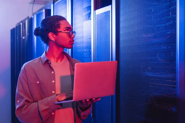 Afrikanisch-amerikanischer Techniker mit Laptop schaut sich Server im Rechenzentrum im Neonlicht an — Stockfoto