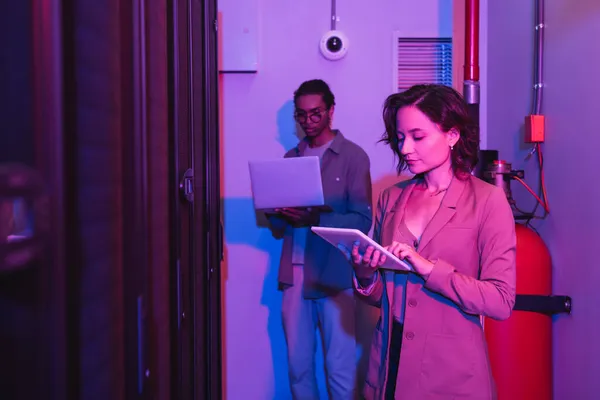 Interracial programmers using gadgets while working in data center in neon light — Stock Photo