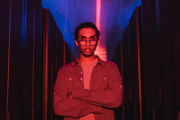 African american programmer looking at camera while standing with crossed arms in data center with neon light — Stock Photo