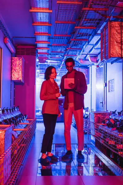 Interracial technicians looking at digital camera while standing in technical equipment room in neon light — Stock Photo