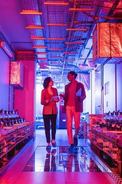 Ingénieurs multiethniques avec des gadgets parler tout en marchant dans la salle d'équipement technique dans le centre de données — Photo de stock