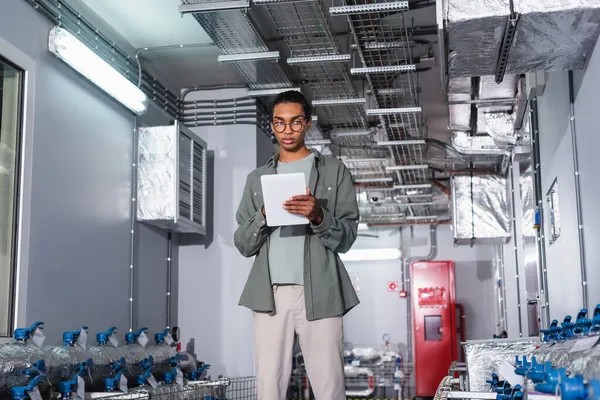 Young afican american engineer using digital tablet while standing near cooling system  of data center — Stock Photo