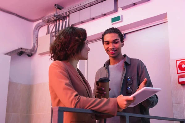 Technicien afro-américain souriant avec tasse en papier pointant vers la tablette numérique tout en travaillant avec un collègue dans un centre de données — Photo de stock