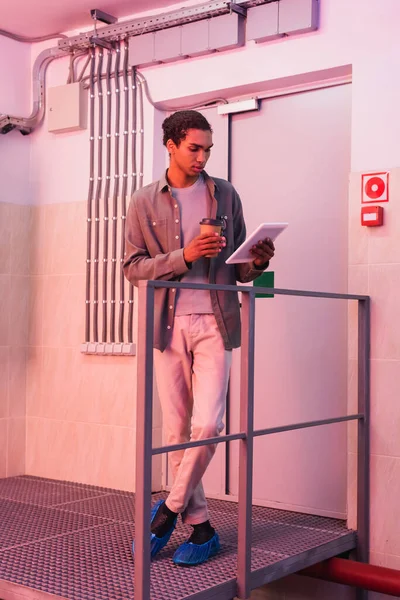 Young afican american engineer looking at digital tablet while standing with takeaway drink in data center — Stock Photo