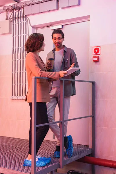 Programmeur afro-américain souriant avec café pour aller pointant vers la tablette numérique près de collègue dans le centre de données — Photo de stock