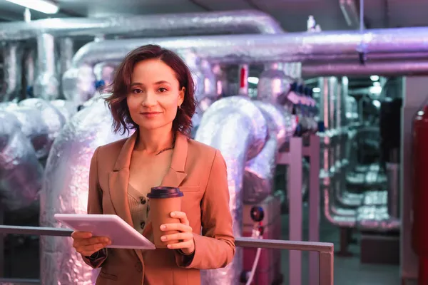 Young engineer with digital tablet and coffee to go smiling at camera in technical room of data center — Stock Photo